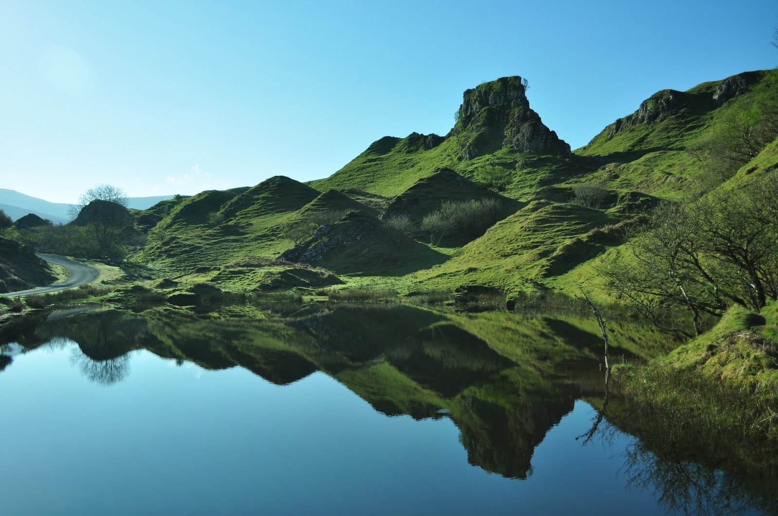 Rolling green hills and a reflective lake under a bright blue sky.